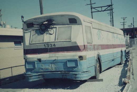 Rear three-quarter view of a white and blue CTA bus, number 1324, parked in a gravel lot with rust, graffiti, and coiled wires on its roof.