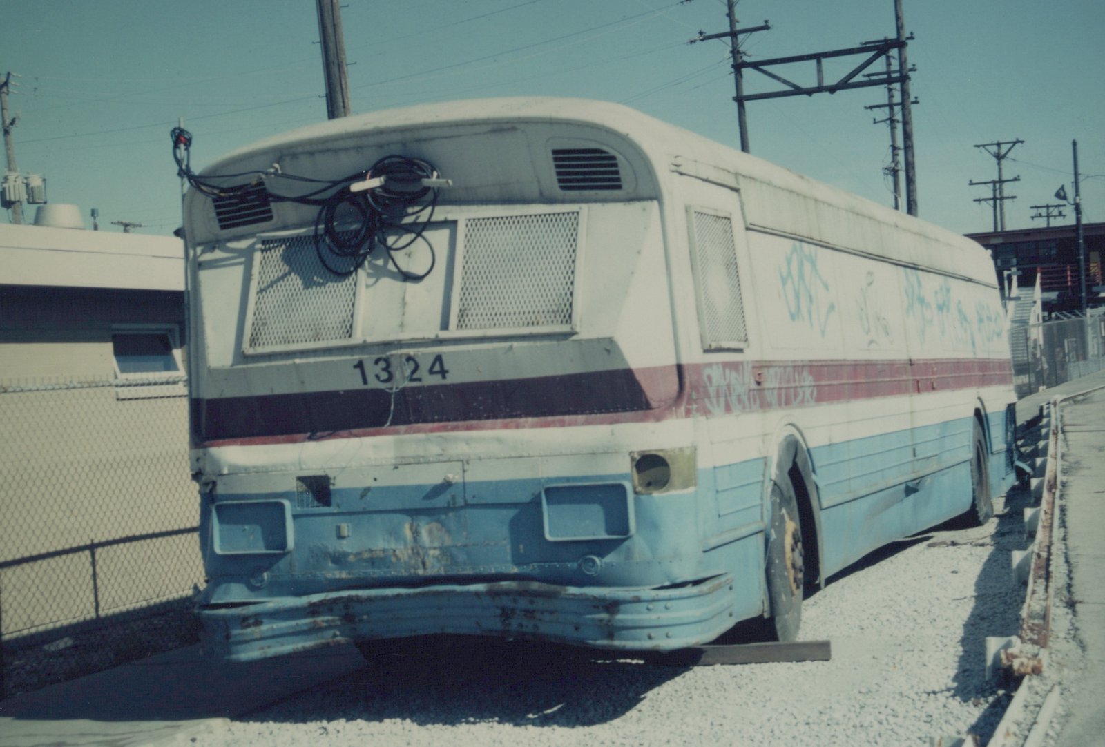Rear three-quarter view of a white and blue CTA bus, number 1324, parked in a gravel lot with rust, graffiti, and coiled wires on its roof.