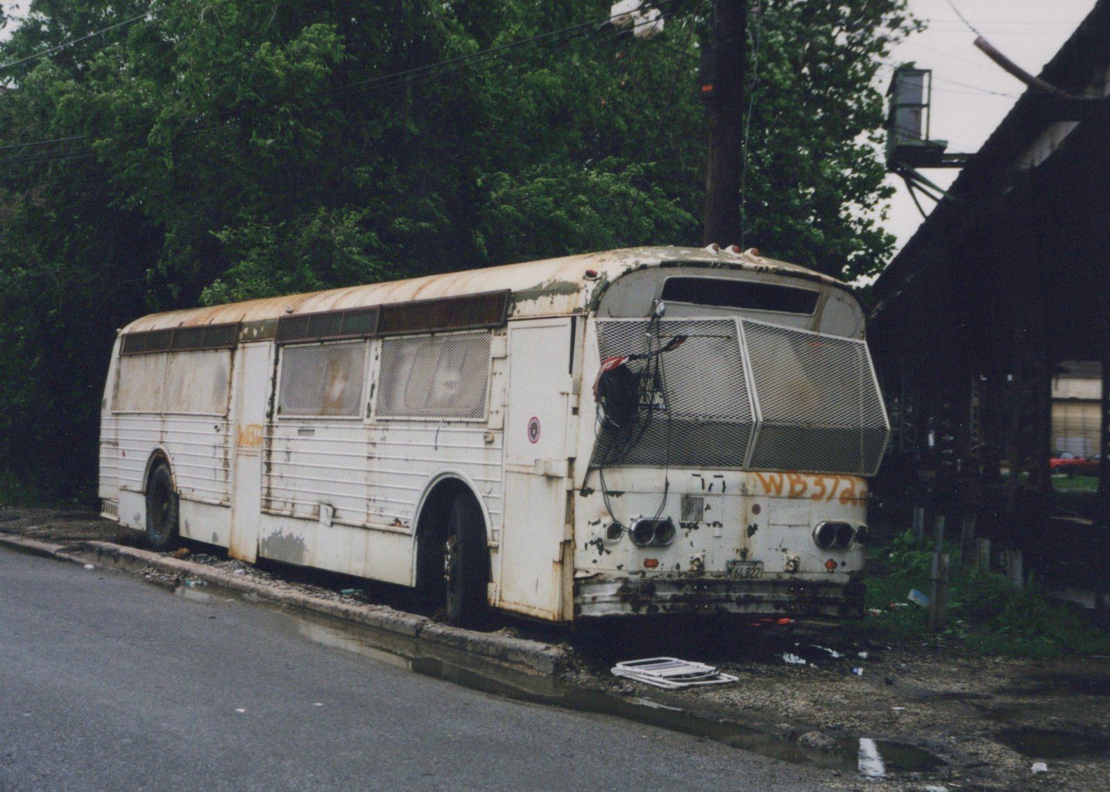 A rusty, white, out-of-service Flxible New Look bus, numbered 717, parked on the side of a wet street with trees and a building in the background.