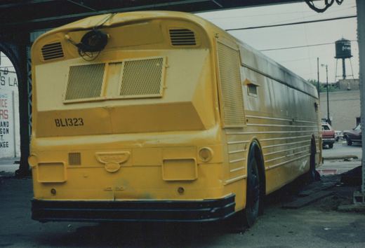 Rear three-quarter view of a yellow Burlington Lines motor bus, fleet number BL1323, parked on a street under a bridge with cars in the background.