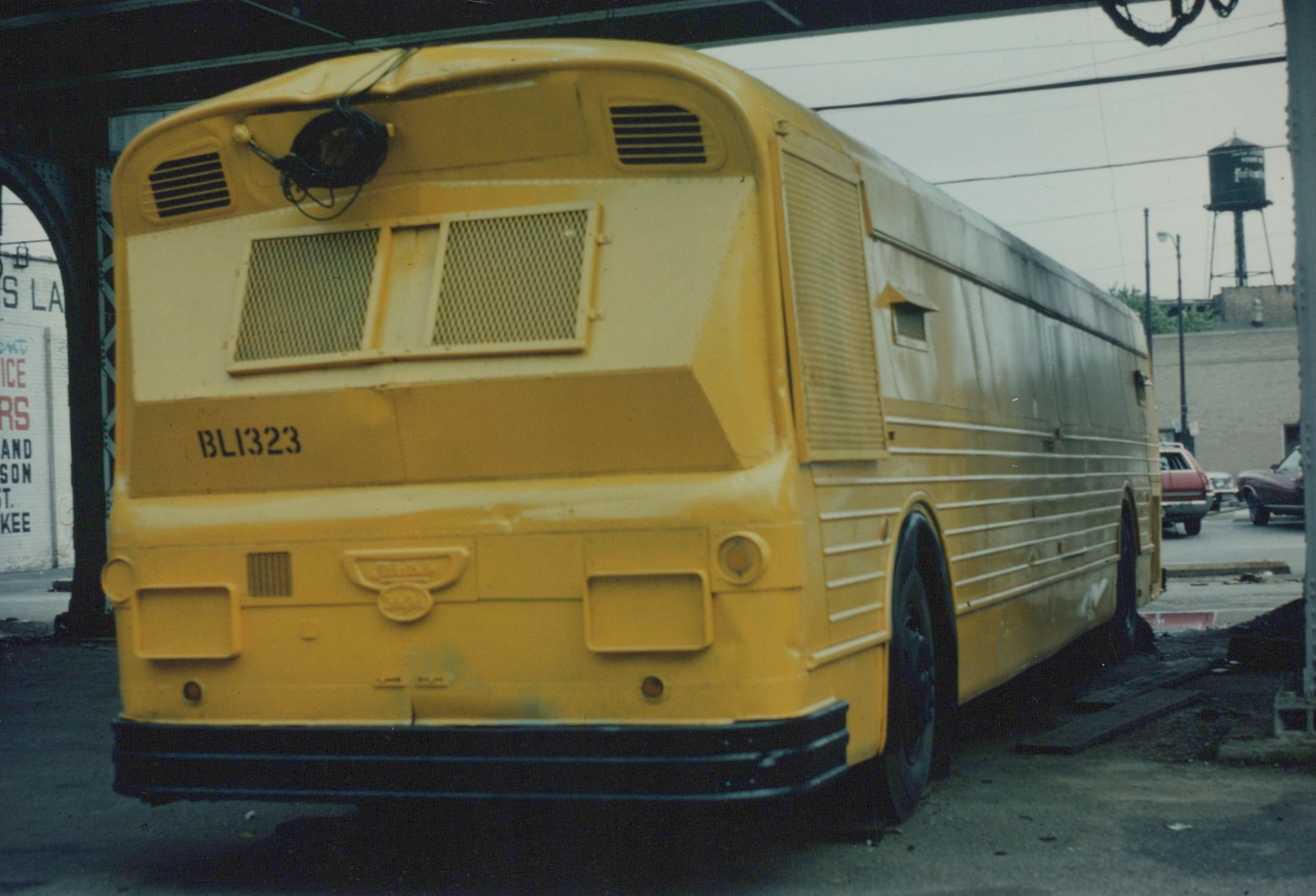 Rear three-quarter view of a yellow Burlington Lines motor bus, fleet number BL1323, parked on a street under a bridge with cars in the background.