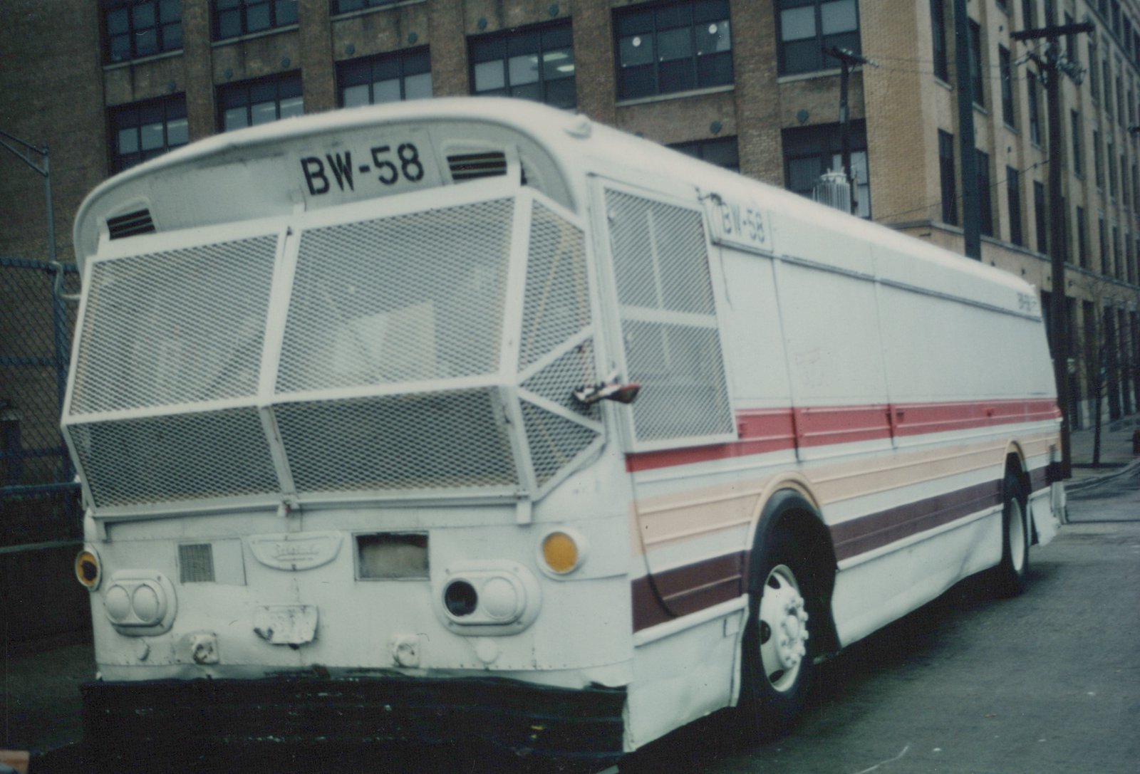 A white Flxible New Look bus with red, peach, and maroon stripes, marked BW-58, parked on a city street with a brick building in the background.