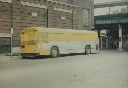 A yellow and white bus with the number BW 56 on its front is parked on a city street next to a brick building, with two people standing under an elevated railway.