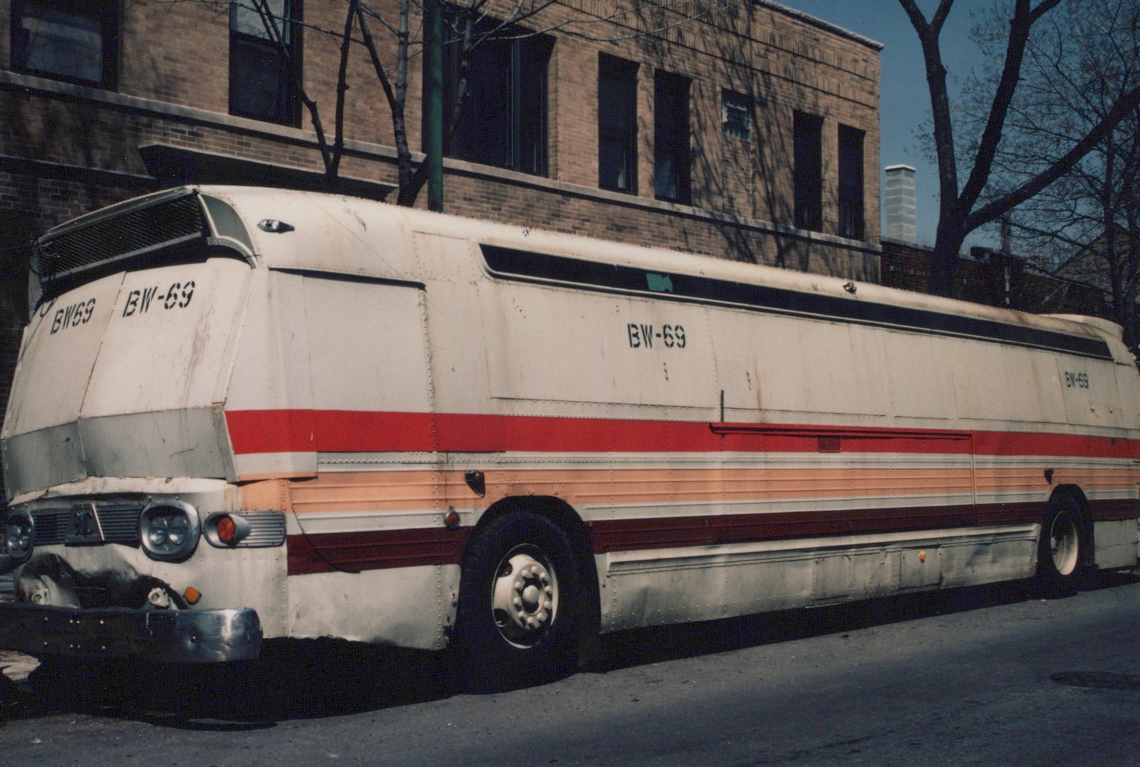 A white and red Flxible Metro motor bus with the fleet number BW-69 is parked on a street next to a brick building, with bare trees in the background.