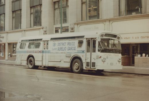 A white Chicago Police motor bus, labeled for the 13th District and Alderman Aurelio Garcia, is parked on a wet city street in front of a building with storefronts.