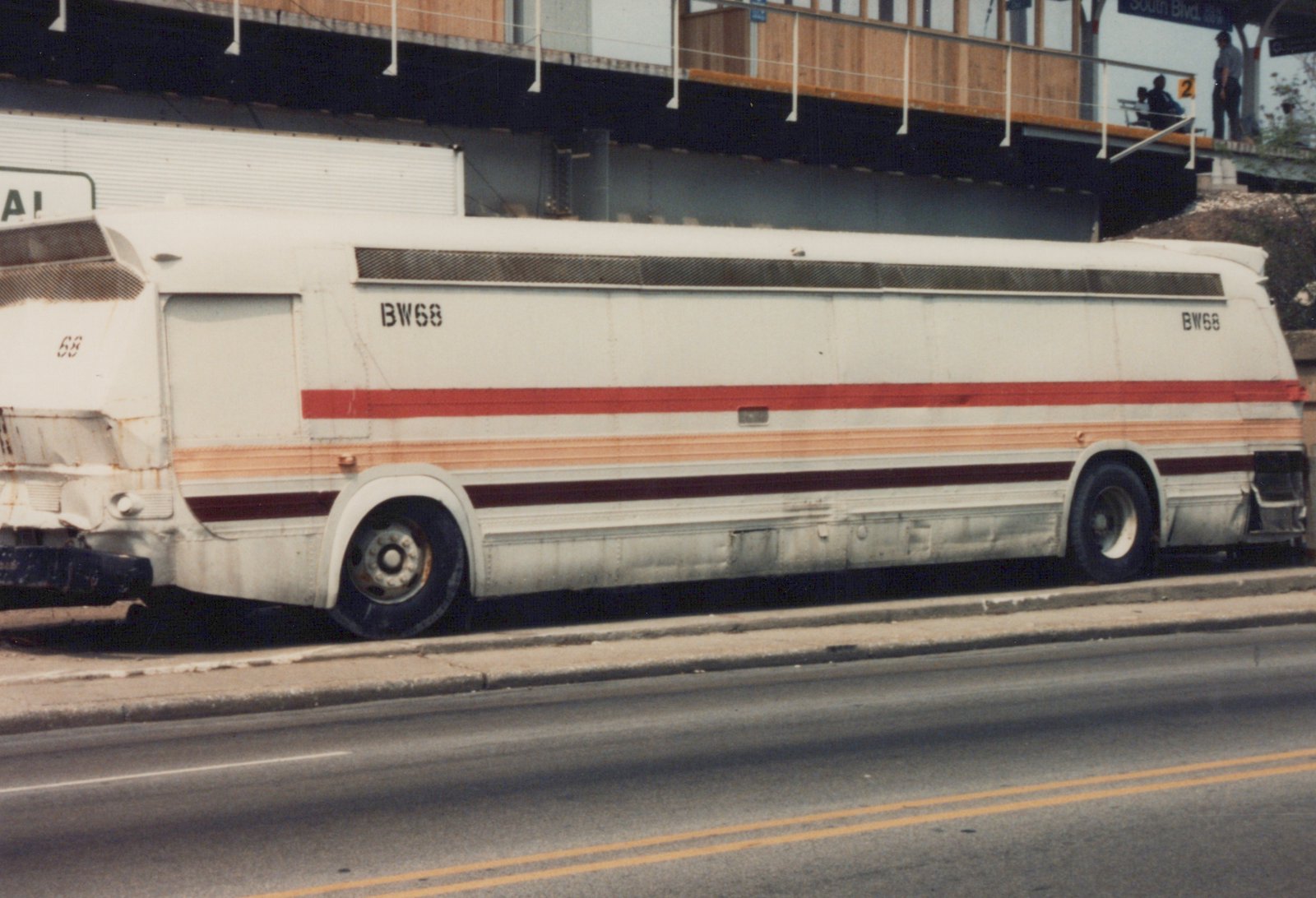 A side view of a white intercity bus with red and orange stripes, marked BW68, parked on a street next to a curb with an elevated structure overhead.