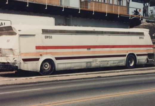 A side view of a white intercity bus with red and orange stripes, marked BW68, parked on a street next to a curb with an elevated structure overhead.
