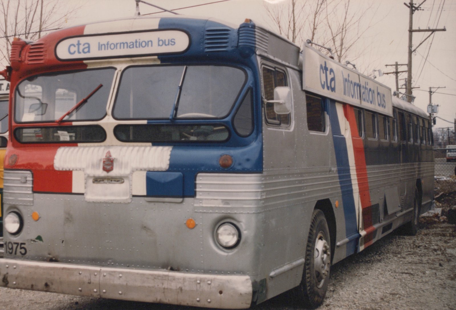 A three-quarter front view of a Chicago Transit Authority bus, number 1975, with 'cta Information bus' signs on the front and side, parked on a gravel lot with bare trees and utility poles in the background.