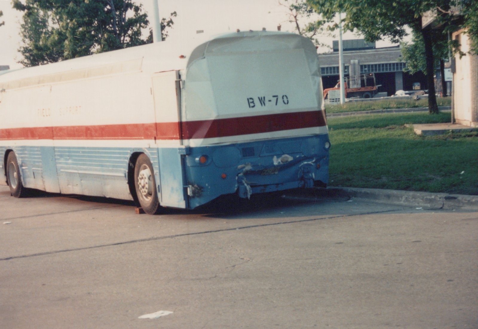 Rear three-quarter view of a white, red, and blue GMC bus with 'BW-70' on the back, parked on a paved lot with green grass and a building in the background.