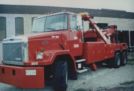 A red AutoCar Top Gun heavy-duty wrecker with fleet number 300 and 'cla' lettering, parked on a gravel surface in front of a white brick building on an overcast day.