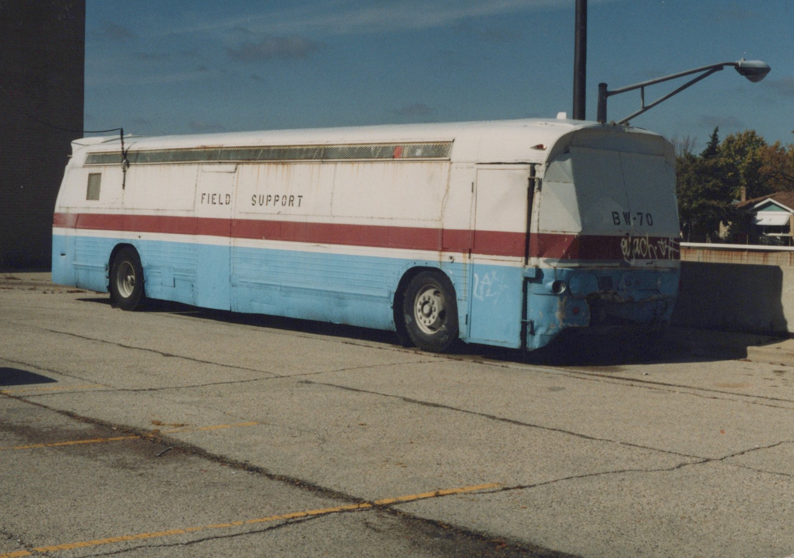 A white, red, and light blue bus, labeled 'FIELD SUPPORT' and 'BW-70' on its rear, is parked on a cracked asphalt lot with yellow painted lines, under a clear blue sky.
