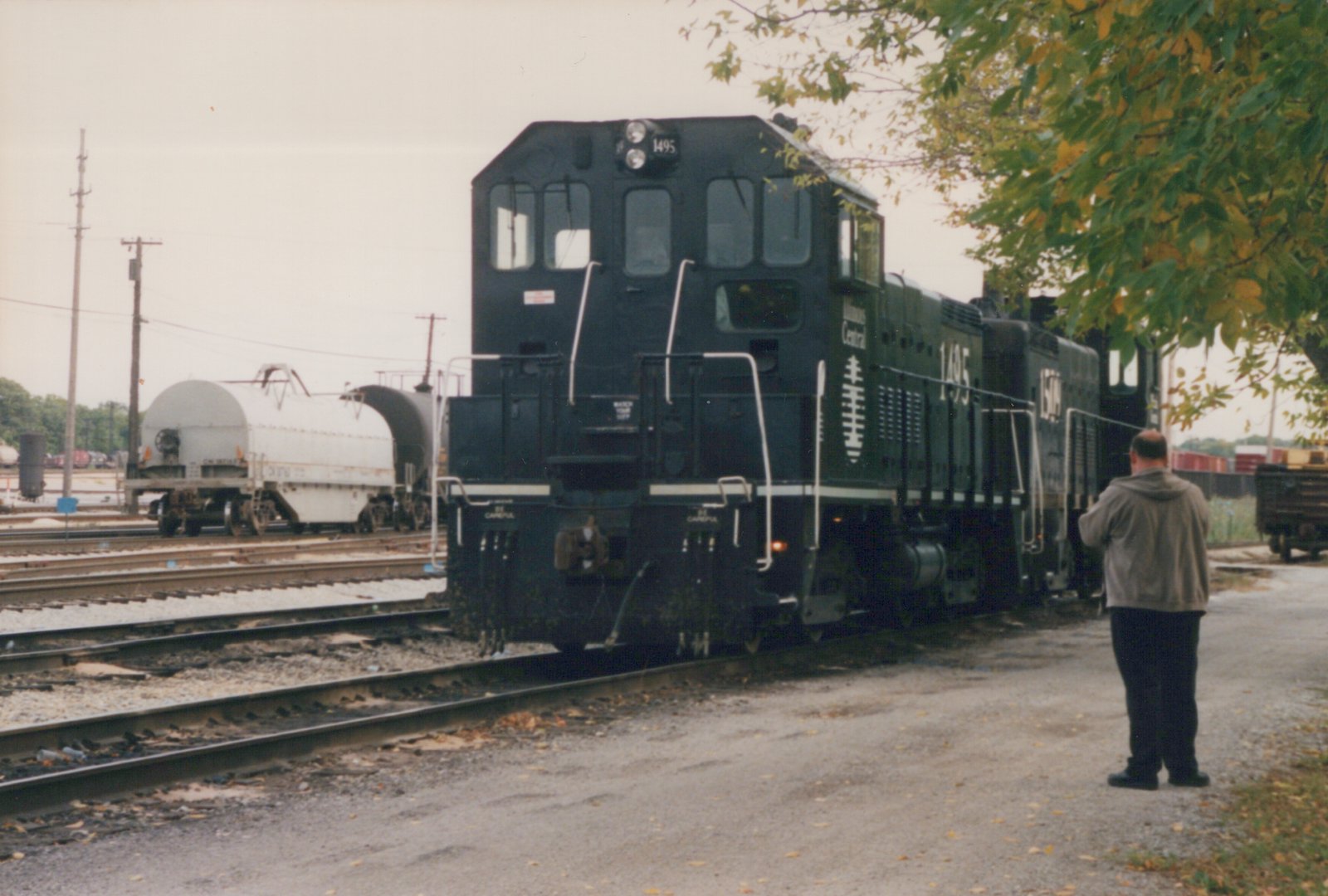 A black Illinois Central switcher numbered 1495, coupled to a second black switcher, stands on a yard track beside a gravel road, with a man photographing it and a gray coil car and a tank car on the tracks at left.