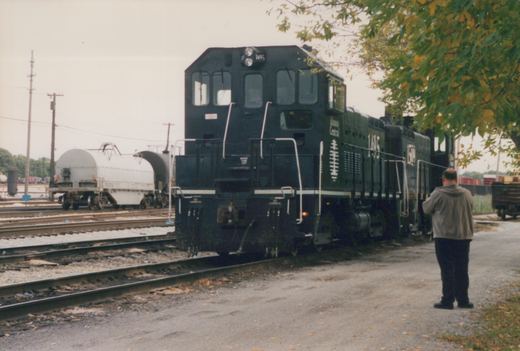 A black Illinois Central switcher numbered 1495, coupled to a second black switcher, stands on a yard track beside a gravel road, with a man photographing it and a gray coil car and a tank car on the tracks at left.