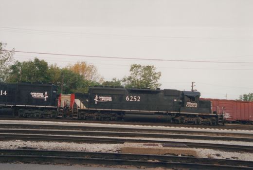 Side view of two black diesel locomotives coupled together with white Operation Lifesaver logos and Illinois Central lettering on the cab of the lead unit, a red boxcar behind, and several tracks in the foreground.
