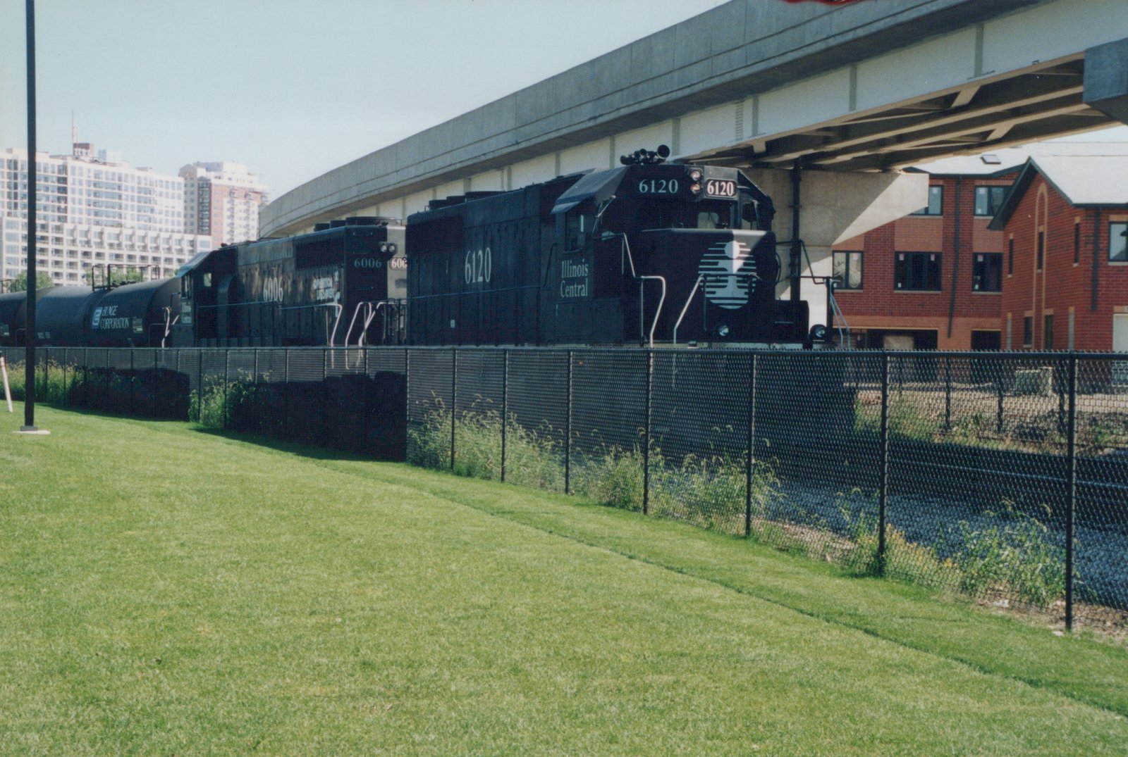 Two black Illinois Central diesel locomotives pulling black tank cars behind a chain-link fence and a lawn, passing under a concrete highway viaduct with high-rise apartments and brick townhouses in the background.