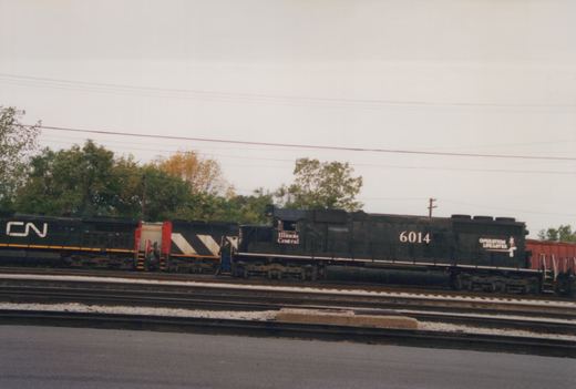 A black Illinois Central diesel locomotive numbered 6014, coupled to two black Canadian National locomotives, on a track in a multi-track yard with trees behind it.