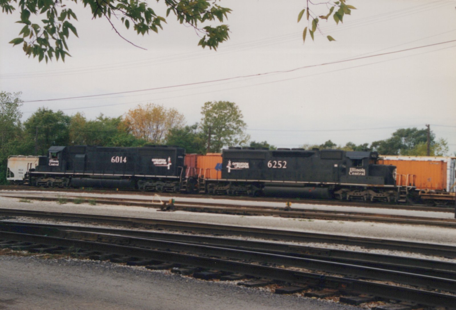 Two black Illinois Central diesel locomotives, numbered 6014 and 6252, with Operation Lifesaver logos stand on a yard track beside a covered hopper and orange containers, with several tracks in the foreground.