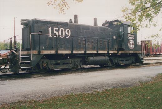 A black diesel switcher with large white numerals 1509 on its hood and Illinois Central lettering on the cab, parked on a track beside a gravel road and grass, with a red flatcar behind it.