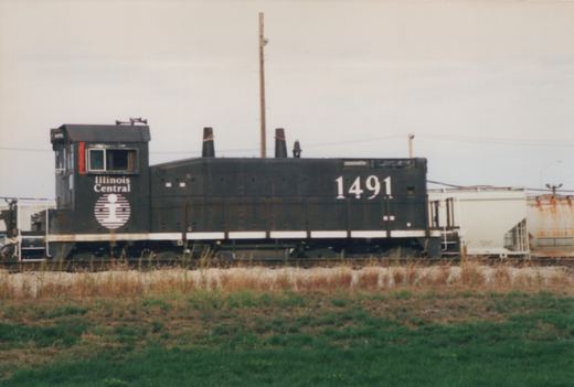 Side view of a black Illinois Central switcher locomotive numbered 1491, with two exhaust stacks and a white stripe, on a track beside grass with white and rusty covered hoppers behind it under an overcast sky.