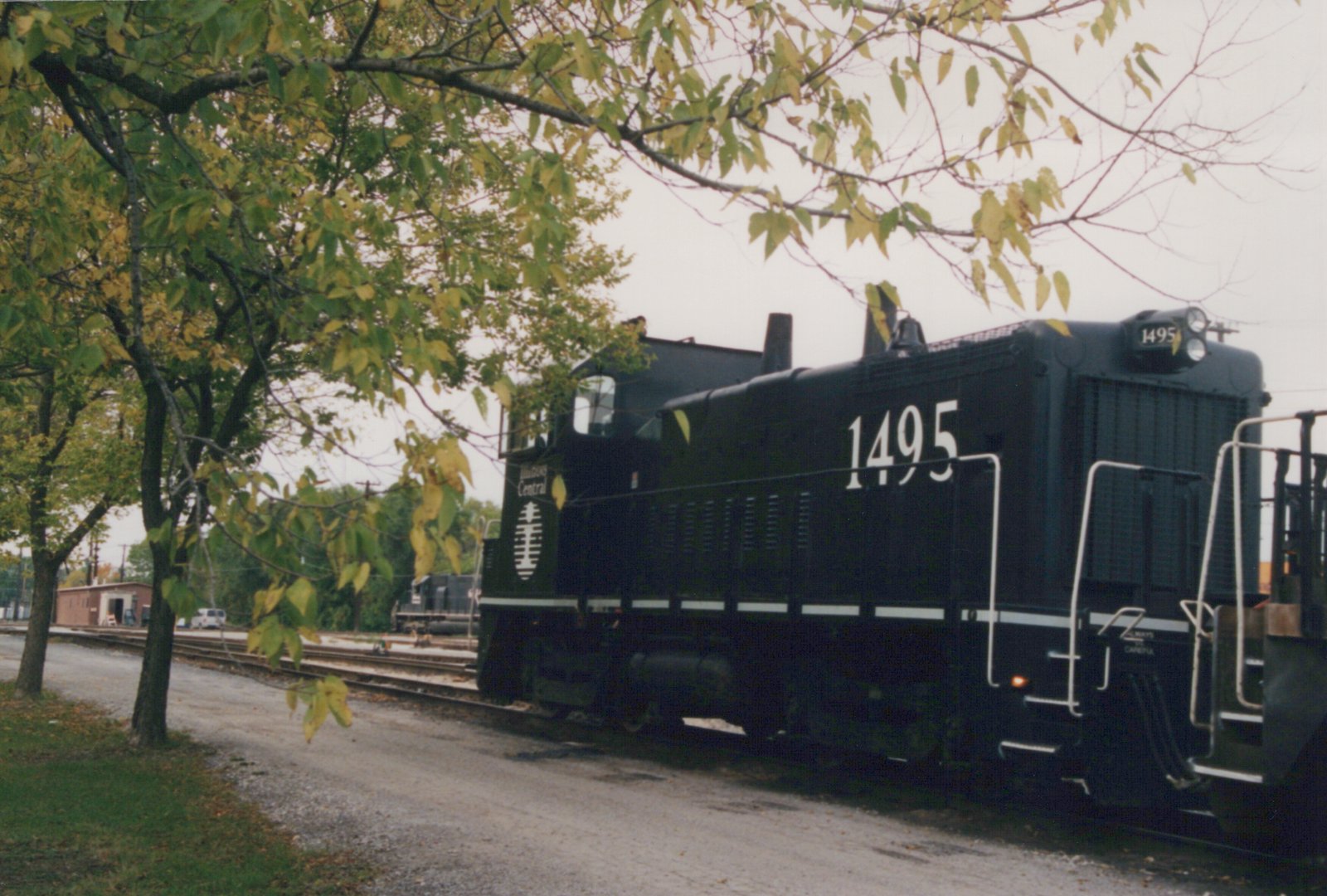 A black diesel switcher numbered 1495 with Illinois Central lettering stands on yard track beside a gravel road, framed by trees with yellowing leaves.