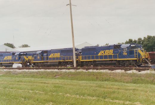A row of dark blue diesel locomotives with yellow railings and large yellow KBS lettering stand on a track behind a grassy field, with a utility pole in front and a white building behind them.
