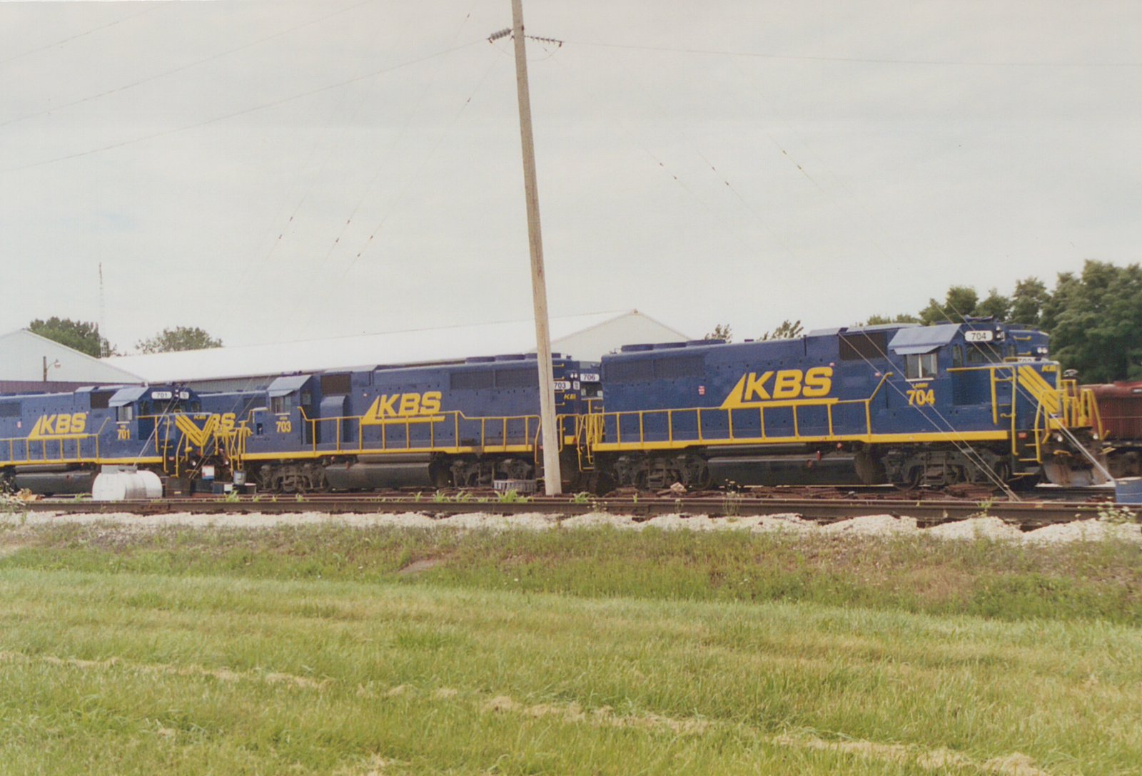 A row of dark blue diesel locomotives with yellow railings and large yellow KBS lettering stand on a track behind a grassy field, with a utility pole in front and a white building behind them.