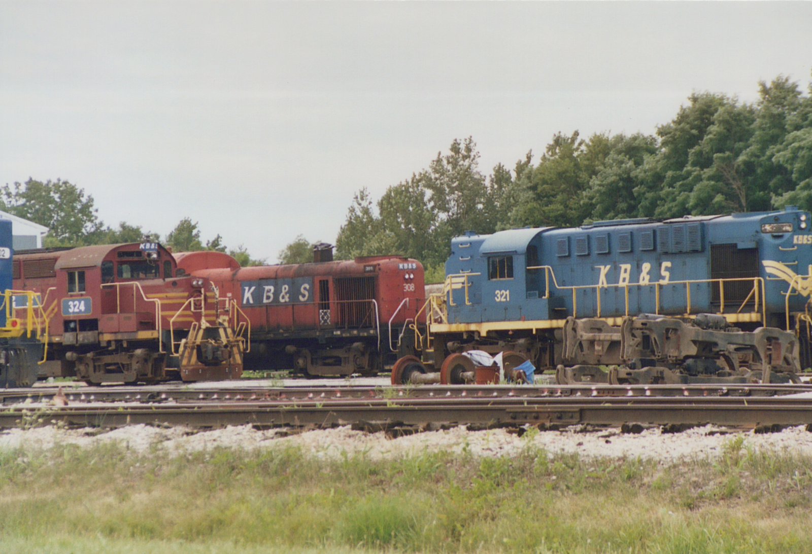 A faded red switcher lettered KB&S and numbered 308 next to a blue KB&S locomotive numbered 321, with loose wheelsets and trucks on the ground beside a yard track and trees behind.
