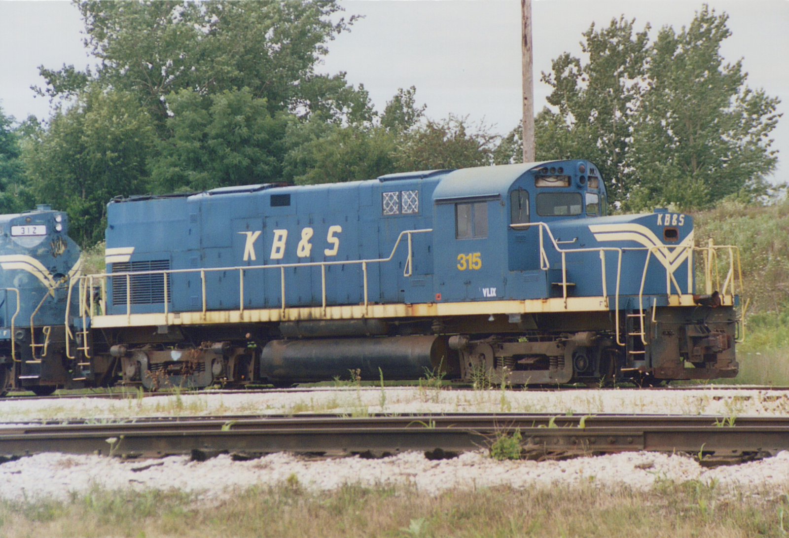 A blue diesel locomotive with cream stripes and K B & S lettering, number 315, parked on a track with trees behind it and the nose of another blue locomotive, number 312, at left.