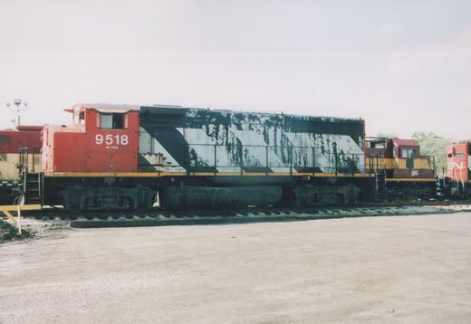A side view of a diesel locomotive with a red cab numbered 9518 and a long hood in peeling black and white diagonal stripes, parked in a gravel yard between a maroon and gold locomotive and a red CN locomotive.
