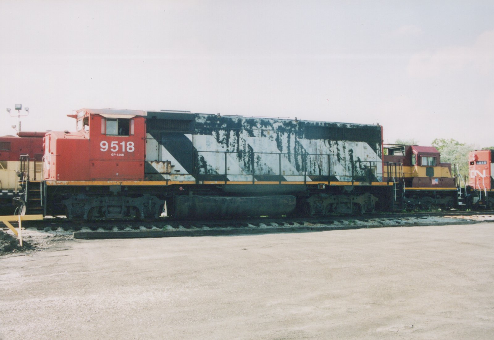 A side view of a diesel locomotive with a red cab numbered 9518 and a long hood in peeling black and white diagonal stripes, parked in a gravel yard between a maroon and gold locomotive and a red CN locomotive.