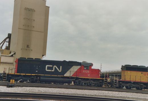 Side view of a black, white and red diesel locomotive lettered CN, numbered 6125 and marked I.C. It is coupled to a yellow and grey locomotive on a yard track, with a tall beige industrial tower behind it under an overcast sky.