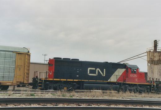 Side view of a black and red Canadian National diesel locomotive numbered 6125, with I.C. under the cab number. It stands between a yellow-ended autorack at left and a rusty sanding tower at right, with tracks in the foreground.