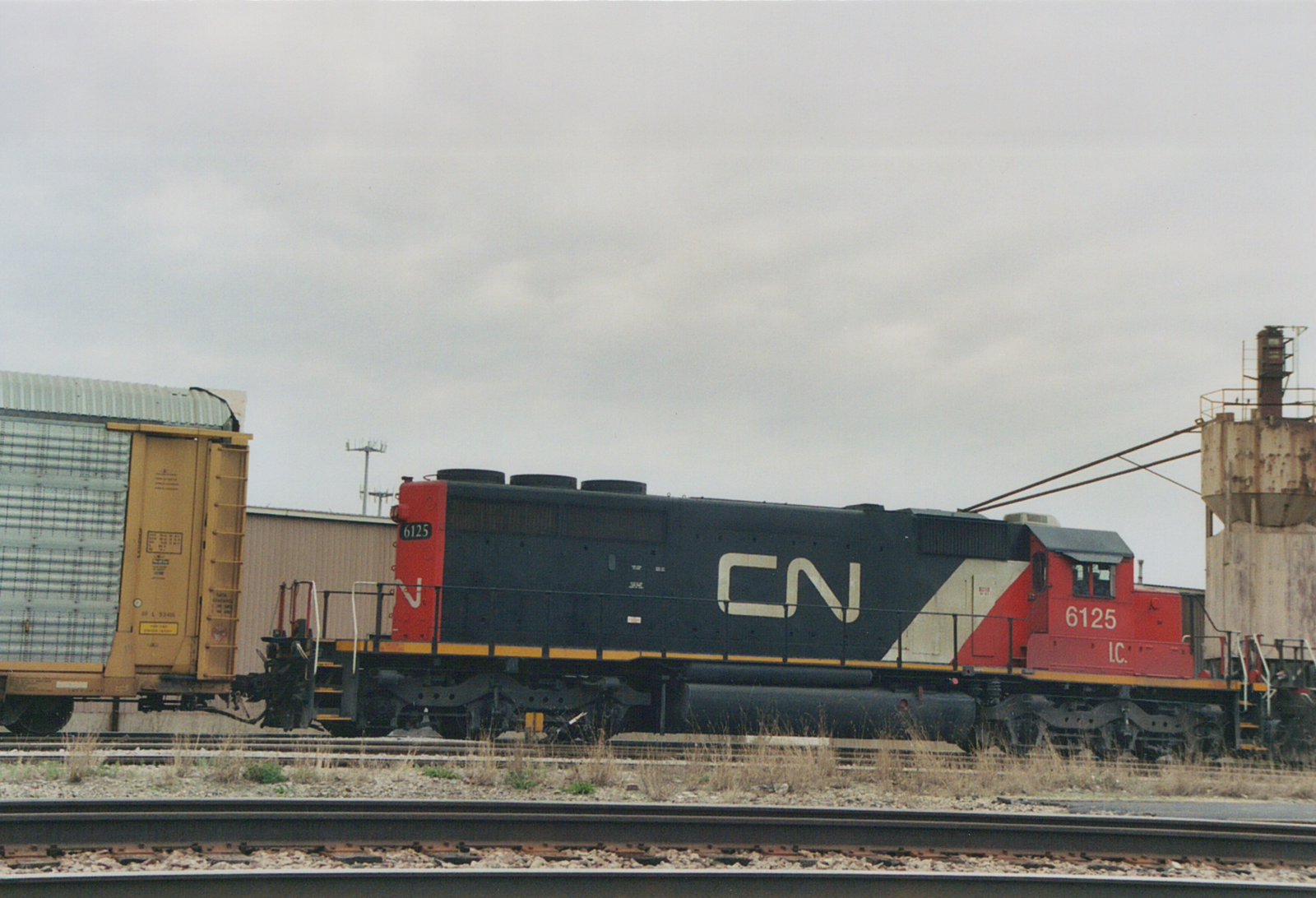 Side view of a black and red Canadian National diesel locomotive numbered 6125, with I.C. under the cab number. It stands between a yellow-ended autorack at left and a rusty sanding tower at right, with tracks in the foreground.