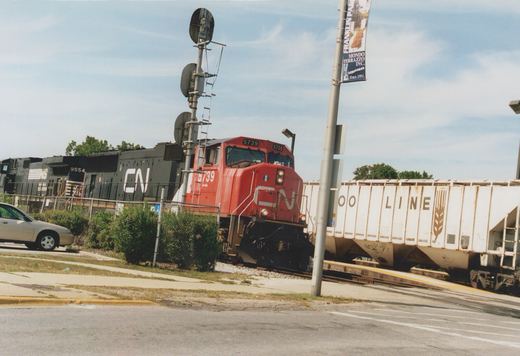 A red CN diesel locomotive numbered 5739, followed by a black CN unit and a black NS unit numbered 9554, approaches a street crossing beside a tall signal mast, with a white Soo Line covered hopper crossing in front and a tan car parked at left.