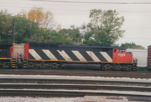 Side view of a red-cabbed locomotive with a black-and-white zebra-striped body on yard tracks, with a covered hopper and another locomotive coupled behind it and trees beyond.