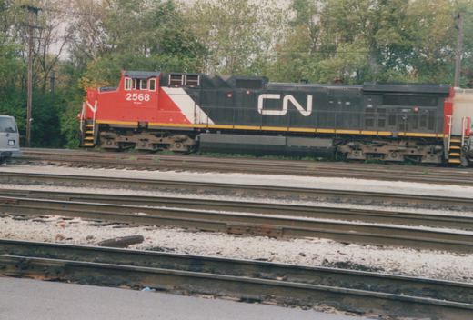 Side view of a red, white and black Canadian National diesel locomotive numbered 2568, with a white CN logo on its black hood, parked beyond several sets of yard tracks with trees behind.
