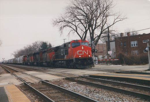 A red and black CN diesel locomotive numbered 2578 leads several more locomotives and a line of gray covered hoppers past a commuter platform and a gated grade crossing, with bare trees and brick apartment buildings behind.