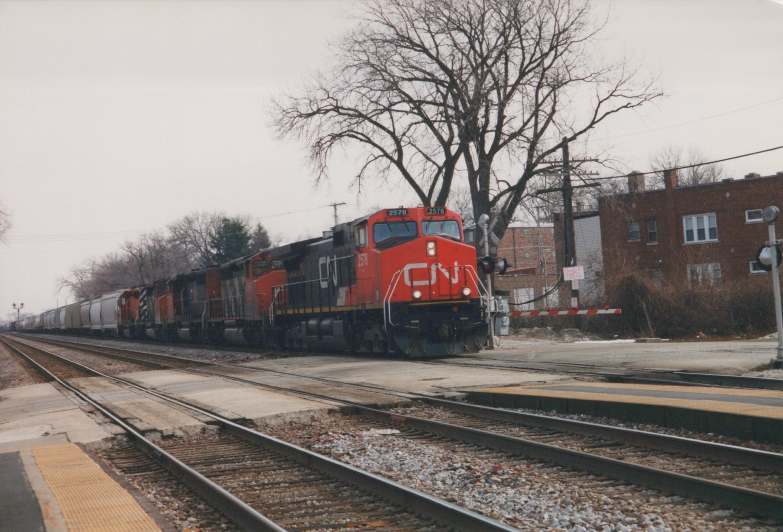 A red and black CN diesel locomotive numbered 2578 leads several more locomotives and a line of gray covered hoppers past a commuter platform and a gated grade crossing, with bare trees and brick apartment buildings behind.