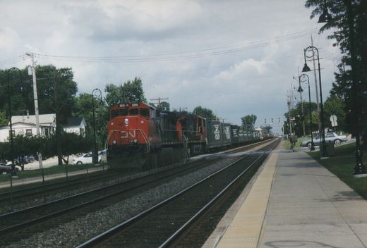 A red and black Canadian National diesel locomotive numbered 2515 leads two more locomotives and a line of green freight cars along a multiple-track main line beside an empty concrete station platform under a cloudy sky.