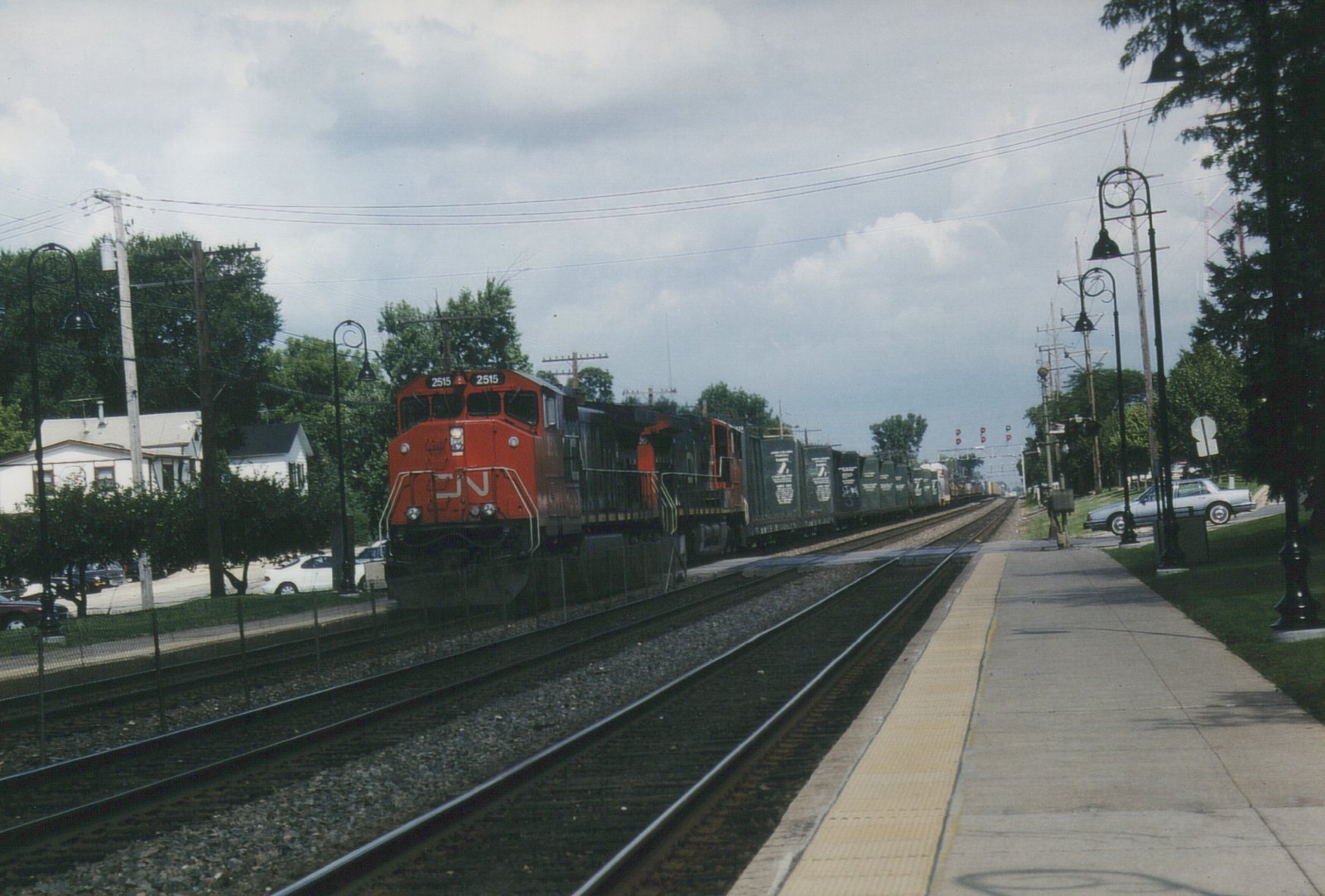 A red and black Canadian National diesel locomotive numbered 2515 leads two more locomotives and a line of green freight cars along a multiple-track main line beside an empty concrete station platform under a cloudy sky.