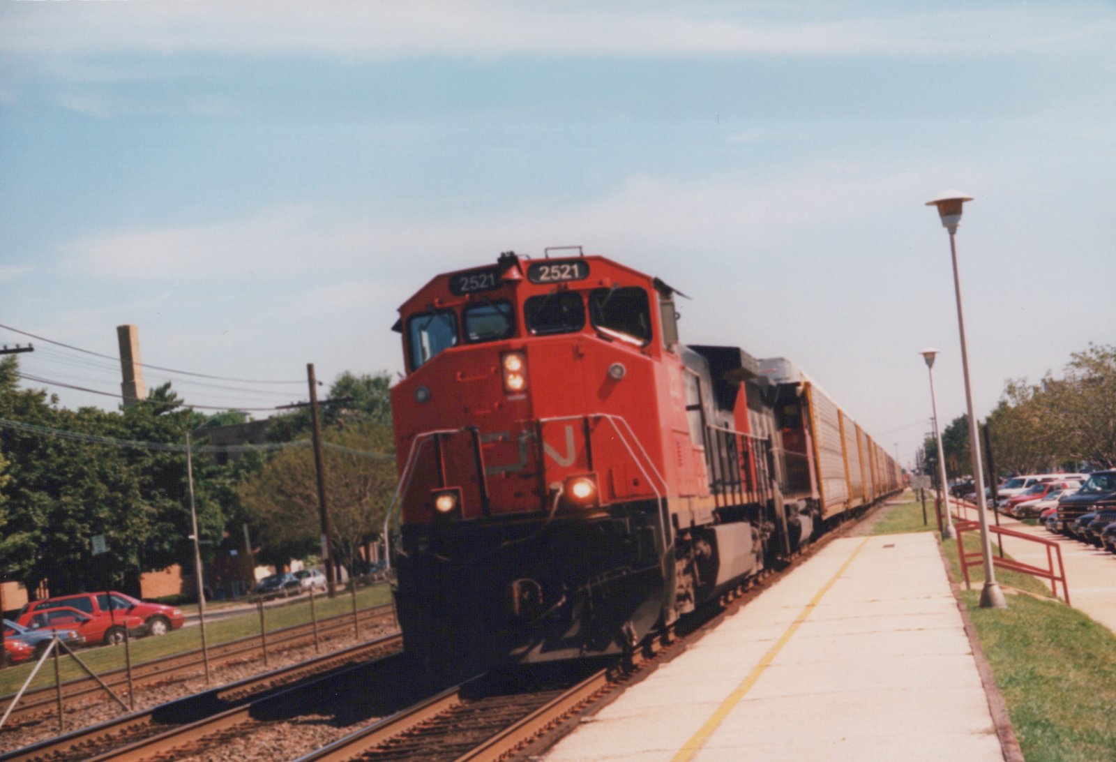 A red-orange Canadian National diesel locomotive numbered 2521, with its headlights on, leads a second locomotive and a long line of white autorack cars past an empty concrete platform with lamp posts, with parked cars on both sides.