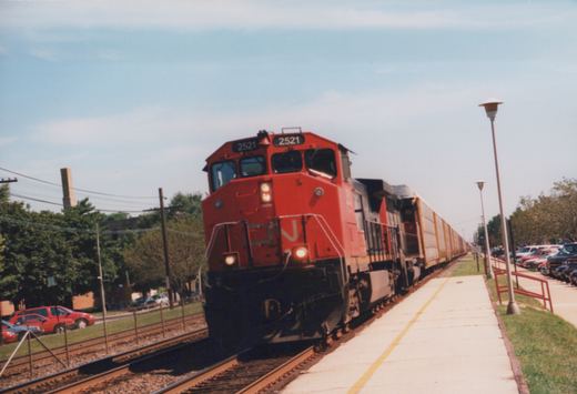 A red-orange Canadian National diesel locomotive numbered 2521, with its headlights on, leads a second locomotive and a long line of white autorack cars past an empty concrete platform with lamp posts, with parked cars on both sides.