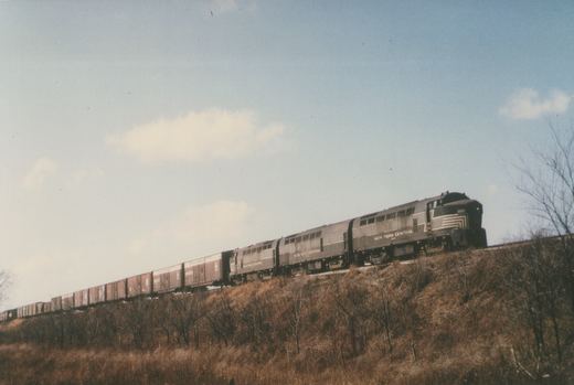Three dark gray New York Central diesel locomotives with a shark-shaped nose and white stripes pull a long line of boxcars along the top of a brushy embankment under a blue sky.