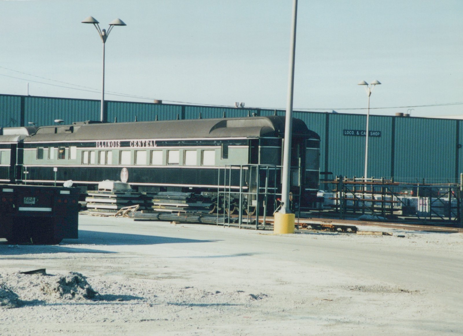 A dark green and light green railroad passenger car lettered ILLINOIS CENTRAL, with an open rear platform, parked beside a large green metal shop building, with stacked materials and a trailer in the foreground.