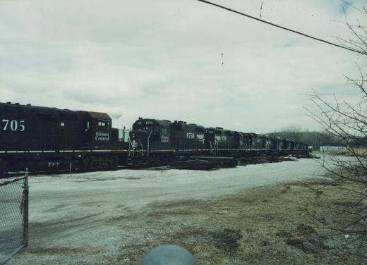Black Illinois Central diesel locomotives numbered 8750 and 8709 parked on a yard track beside piles of ties, with a gravel lot, bare trees and a water tower under an overcast sky.