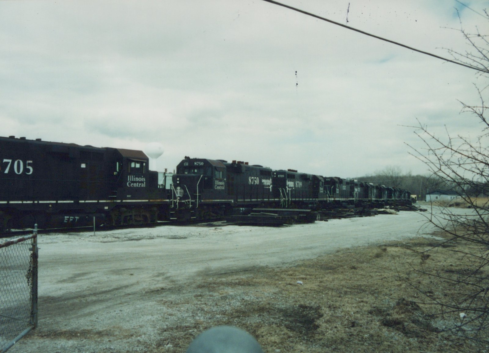 Black Illinois Central diesel locomotives numbered 8750 and 8709 parked on a yard track beside piles of ties, with a gravel lot, bare trees and a water tower under an overcast sky.