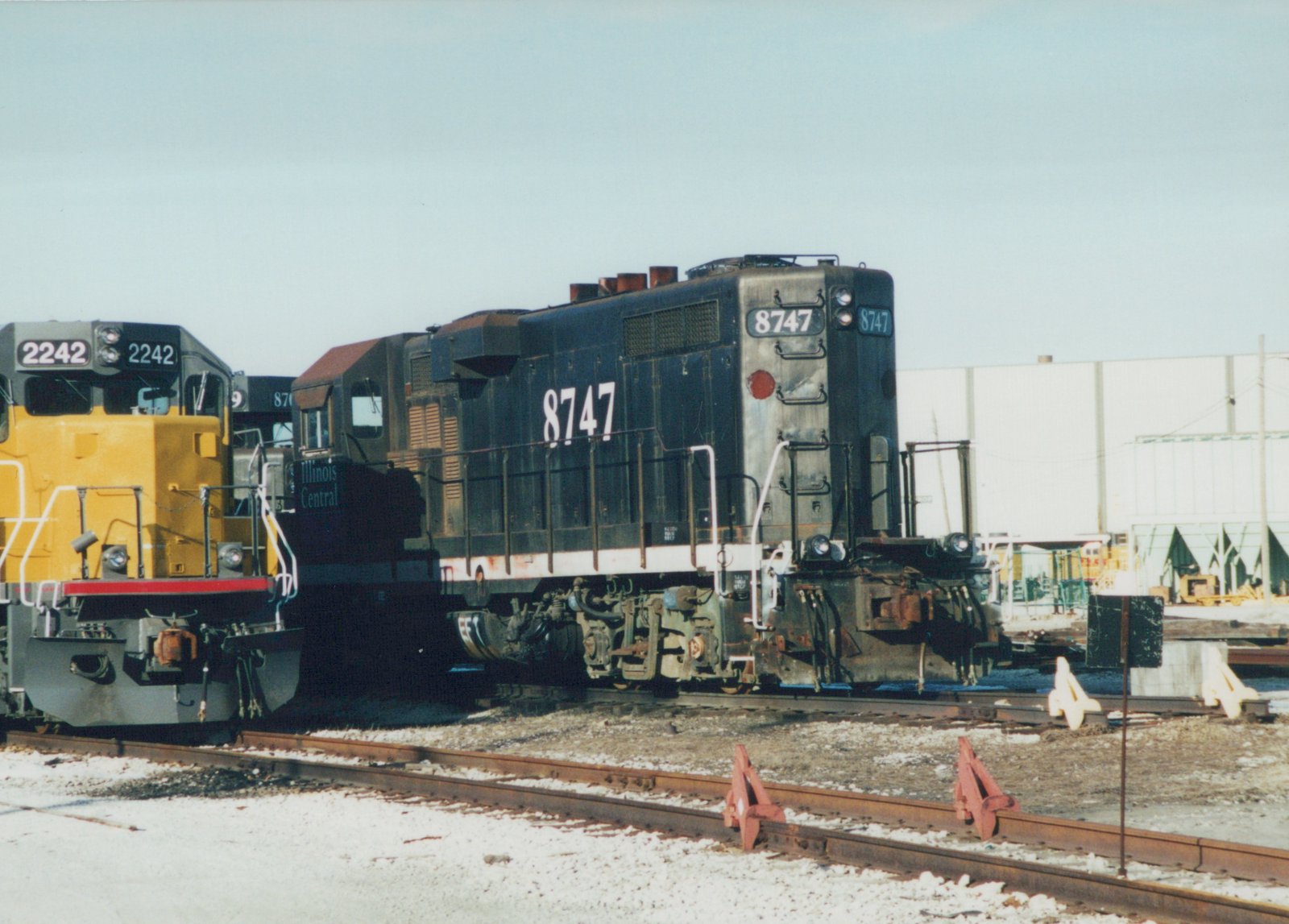 A black Illinois Central diesel locomotive numbered 8747 stands on a yard track beside a yellow and gray locomotive numbered 2242, with red derails on the rails in front.