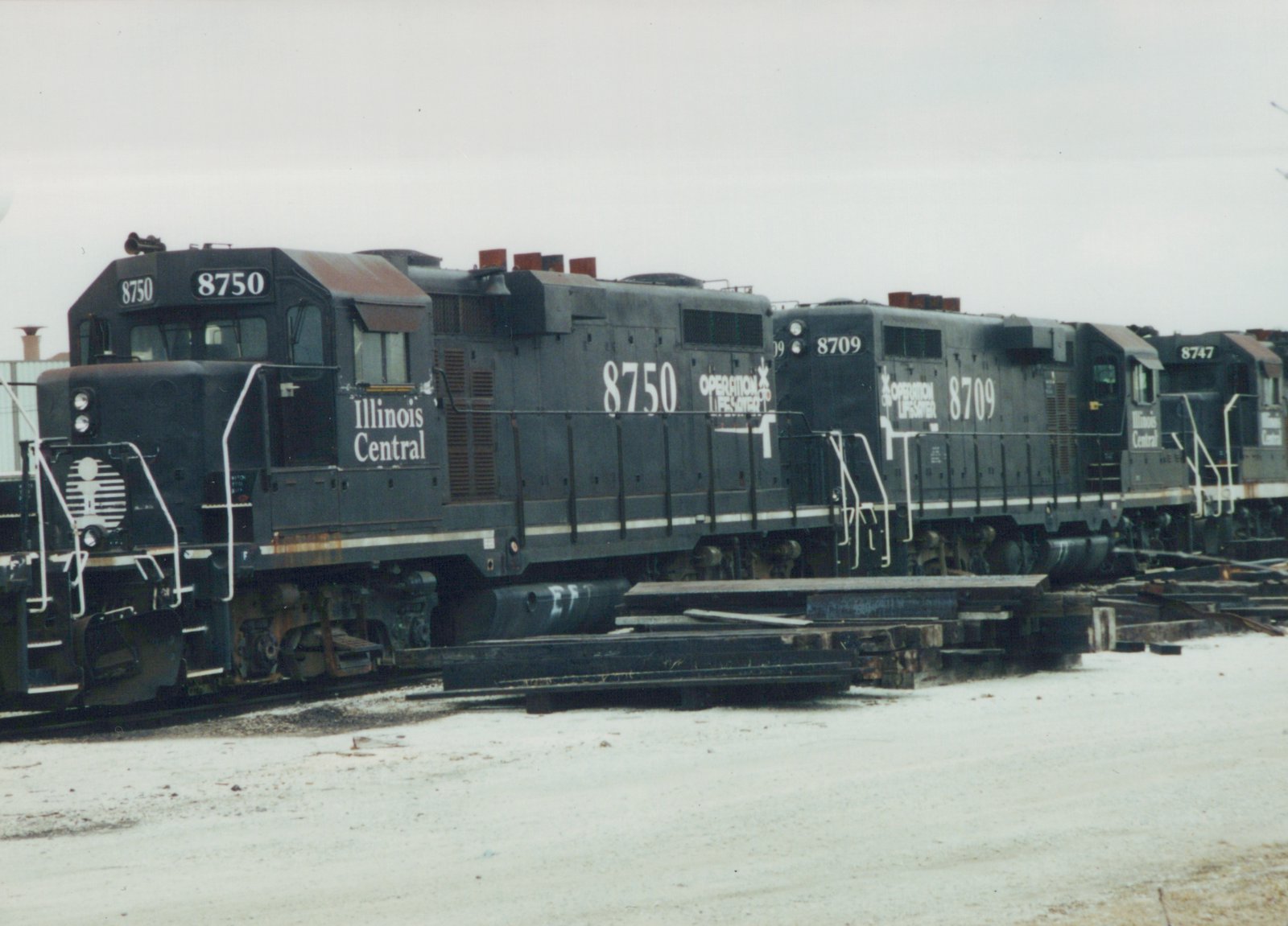 Three black Illinois Central diesel locomotives with white lettering, numbered 8750, 8709 and 8747, stand coupled on a track beside stacks of rail and timbers under an overcast sky.