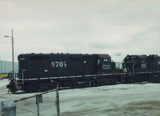 Two black Illinois Central diesel locomotives numbered 8705 and 8750 stand side by side on yard tracks, with a third black locomotive partly in view at right, a chain-link fence in front and a white water tower in the distance.