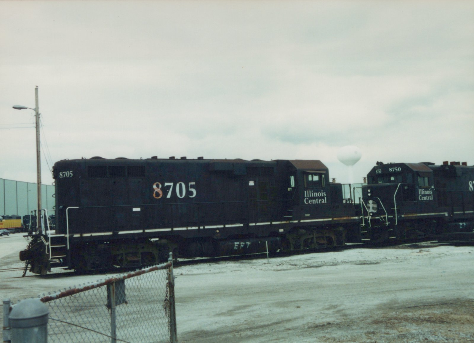 Two black Illinois Central diesel locomotives numbered 8705 and 8750 stand side by side on yard tracks, with a third black locomotive partly in view at right, a chain-link fence in front and a white water tower in the distance.